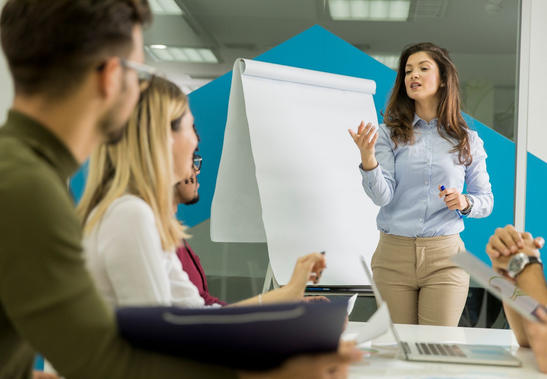 Confident young team leader giving a presentation to a group of young colleagues as they sit grouped by the flip chart in the small startup office Confident young team leader giving a presentation to a group of young colleagues as they sit grouped by the flip chart in the small startup office